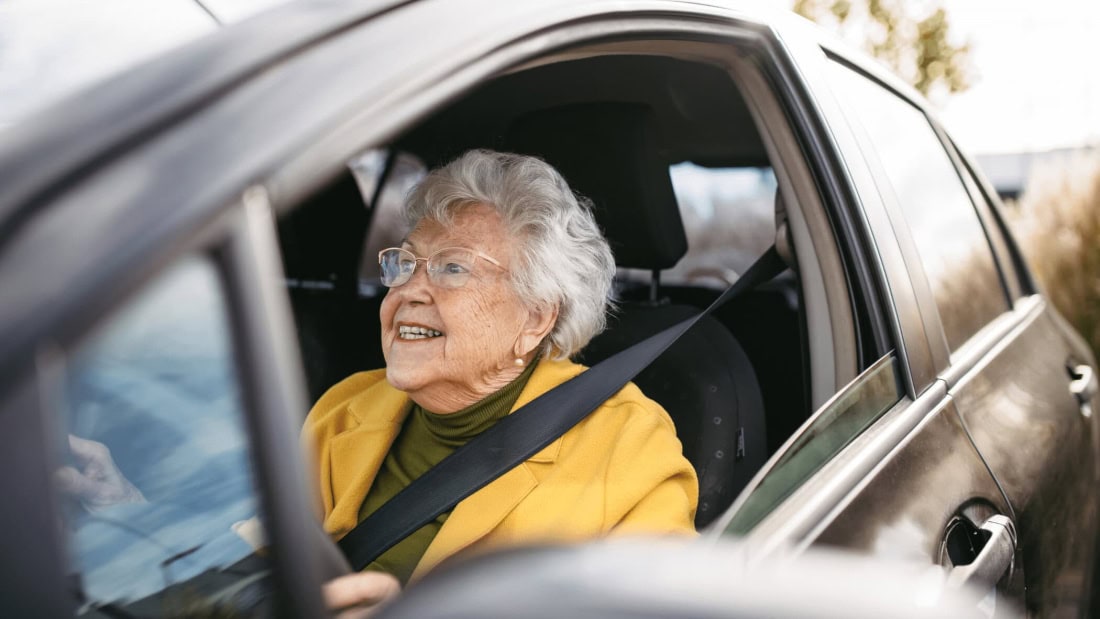 Oudere vrouw in auto met gordel, glimlachend en klaar voor rijbewijskeuring.